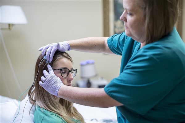 A technician wearing blue scrubs and purple gloves carefully attaches electrodes to a patient's head for a sleep monitoring session