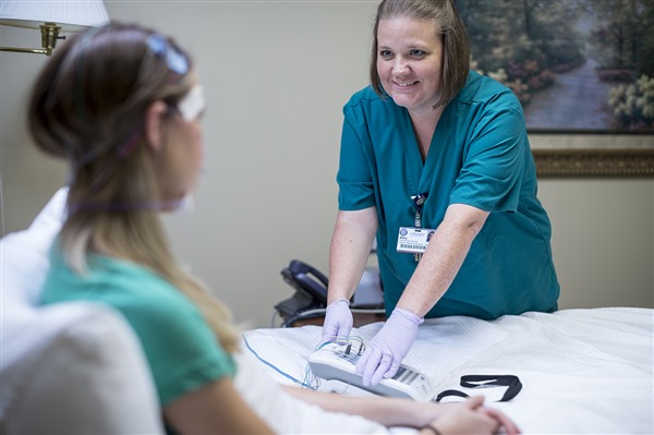 A smiling sleep technician in blue scrubs monitors a seated patient who has electrodes attached to her forehead and temples