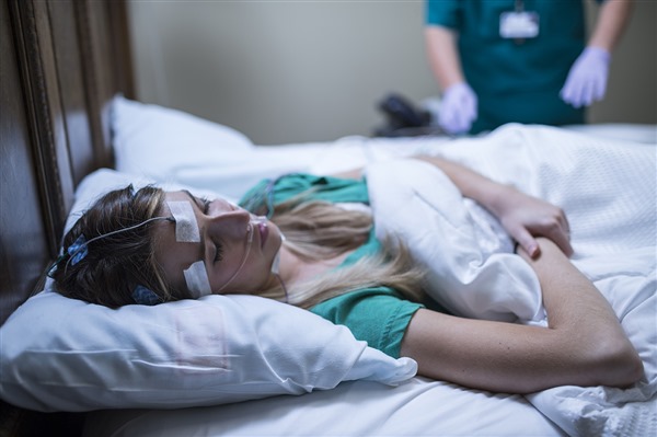 A young woman lies in a bed with several sensors taped to her face and head while a technician works in the background