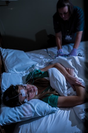 A patient sleeps in a darkened room with various monitoring wires attached to her head as a technician adjusts the equipment